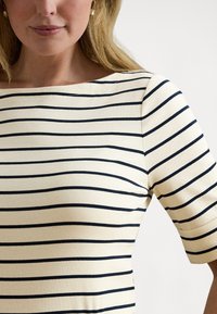 Blondīne sieviete, tērpusies krēmkrāsas kreklā ar melnām horizontālām svītrām, redzama no kakla līdz viduklim baltā fonā.