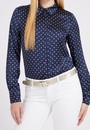 Kobieta ubrana w granatową koszulę w białe kropki, zapinaną na guziki, włożoną do białych spodni z jasnym paskiem z złotą klamrą.