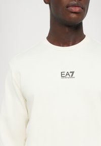Camisa blanca de manga larga hecha de tela suave, con un pequeño logo negro "EA7 Emporio Armani" en el pecho. Cuello redondo, ajuste casual.