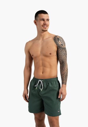 Homme torse nu avec un short de bain vert foncé et une manche de tatouage tribal noire détaillée sur son bras droit, souriant et regardant vers la droite.