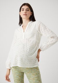 Hvit bomullsbluse med broderte detaljer, krage, og lange, elastiske ermer. Kombinert med grønne mønstrede bukser.