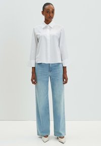 White collared shirt with long sleeves, paired with light blue wide-leg jeans. The jeans have a faded finish, and the outfit includes pointed white shoes.