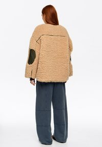 Persona con cabello largo y rojo que lleva una chaqueta beige esponjosa con parches oscuros en los codos y pantalones azul oscuro holgados, de pie de espaldas.