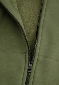 Oliwkowa kurtka zapinana na zamek, wykonana z połączenia gładkiego materiału i puszystego polaru wewnętrznego, z czarnym zamkiem błyskawicznym i detalami w czarnym kolorze.