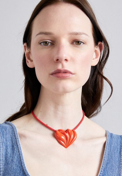 Colgante en forma de corazón rojo hecho de un material brillante, con un diseño texturizado y en espiral, sujeto a un delgado cordón rojo alrededor del cuello.