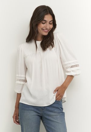 Weiße Bluse mit gefälteltem Vorderteil und Dreiviertelärmeln, verziert mit Spitzenelementen. Getragen über hellblauen Jeans, mit einer weichen, fließenden Textur.