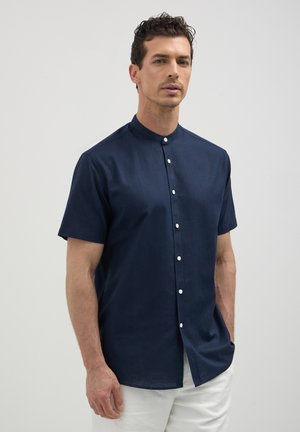 Camicia blu navy a maniche corte con colletto alla mandarino, fronte con bottoni e texture liscia. I bottoni bianchi creano un contrasto con il tessuto scuro.