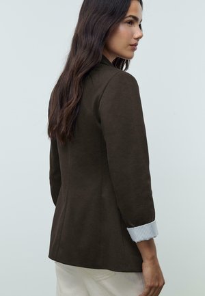 Giacca marrone aderente con maniche a tre quarti, polsini a coste di colore chiaro e una texture liscia, vista da dietro.