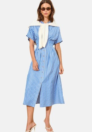 Naine kannab sinist ja valget vertikaalsete triipudega nööpidega midi-kleiti, koorevärvi kampsunit õlgadel, päikeseprille ja valgeid kontsakingi.