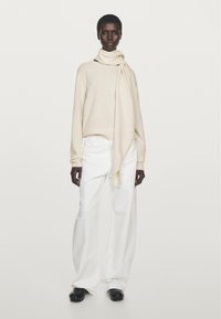 Person wearing a cream sweater with a matching long scarf, wide white pants, and black clogs, standing against a plain light background.