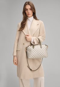 Bolso beige con exterior estampado, herrajes dorados, dos asas y una correa ajustable de color verde oliva, sostenido por una persona con un abrigo beige.
