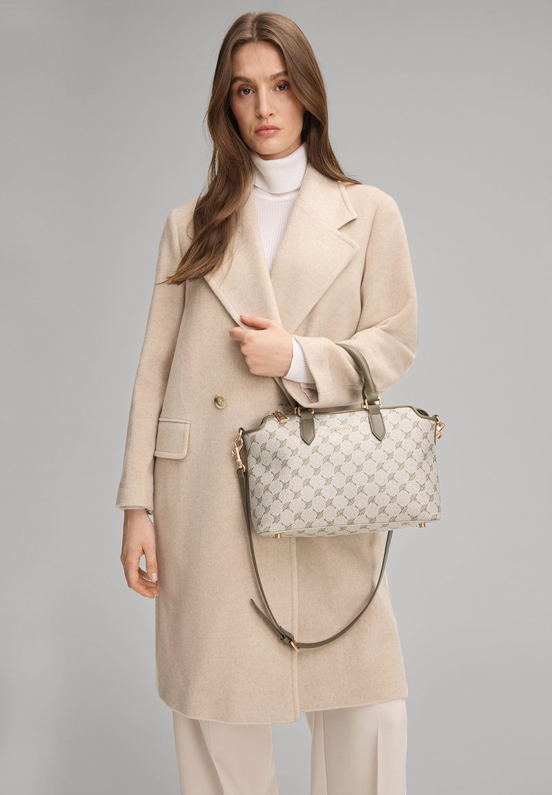 Bolso beige con exterior estampado, herrajes dorados, dos asas y una correa ajustable de color verde oliva, sostenido por una persona con un abrigo beige.