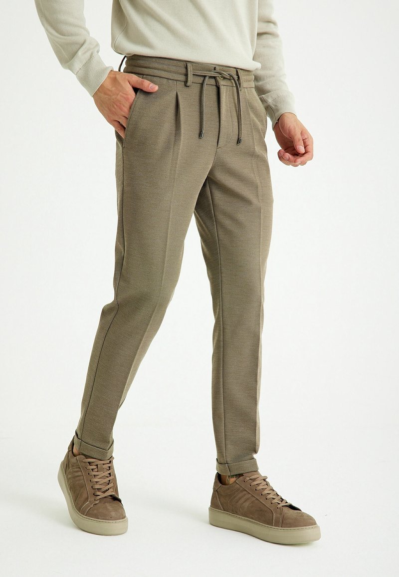 Homme portant un pantalon beige fuselé avec taille à cordon et des baskets en daim marron, debout avec une main dans la poche.