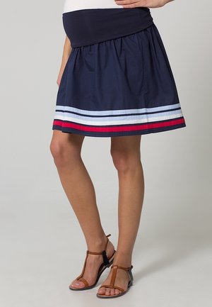 Navyblauwe zwangerschapsrok met witte, lichtblauwe en rode strepen bij de zoom, gedragen met bruine sandalen met bandjes op een lichte achtergrond.