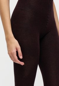 Gros plan sur la main d'une personne près de sa partie supérieure de la cuisse portant des leggings en maille ajustés marron foncé sur un fond clair uni.