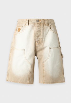 Homme portant un pull bicolore marron et crème, un short beige longueur genou, des chaussettes blanches et des mocassins marron foncé, debout devant un fond blanc.