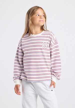 Meisje met lang haar, gekleed in een wit met rood gestreept shirt met lange mouwen met daarop "89" en lichtgrijze broek, kijkend opzij.
