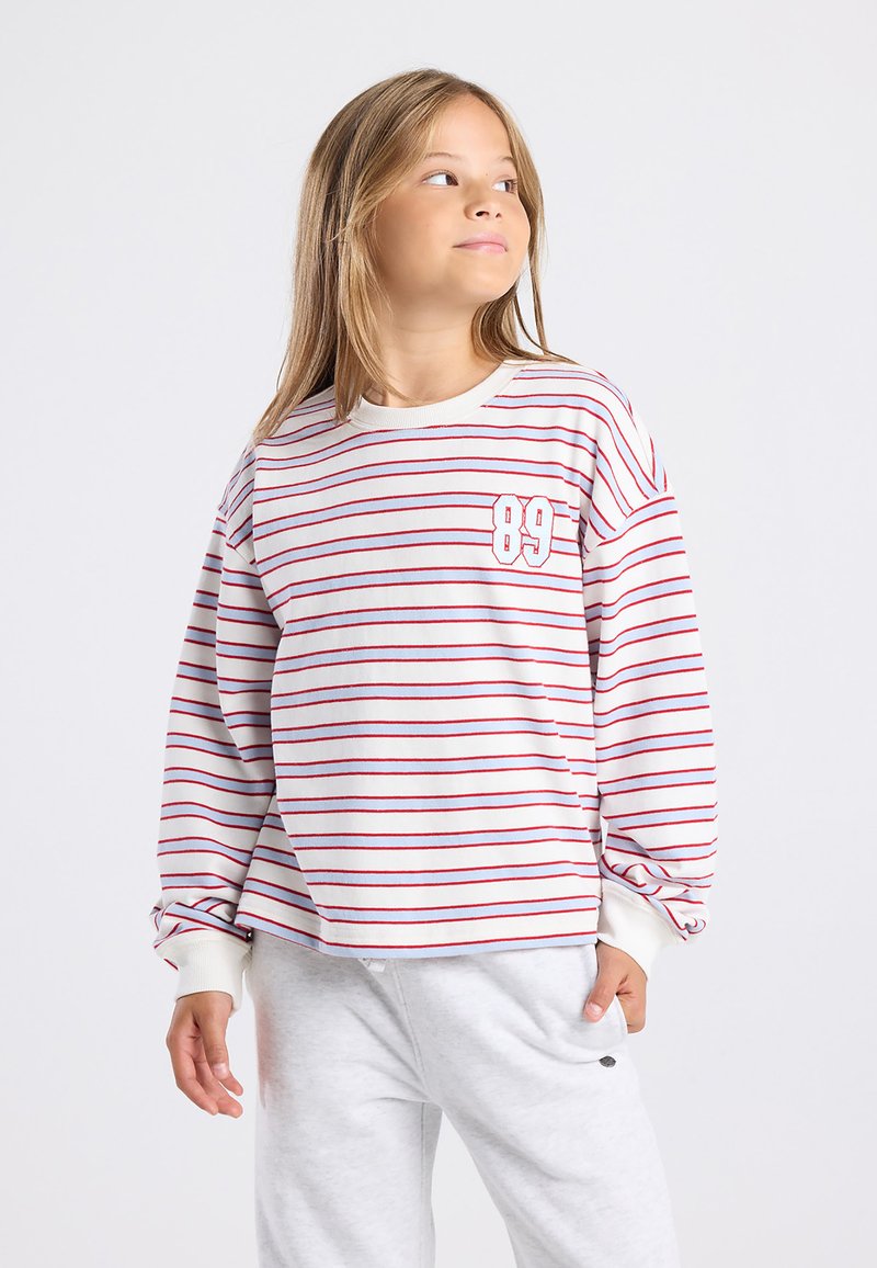 Girl with long hair wearing a white and red striped long-sleeve shirt with "89" and light gray pants, looking to the side.