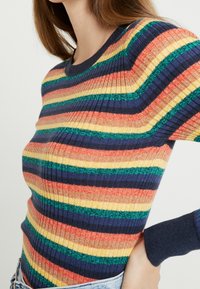 Raidallinen neulepusero, jossa leveät vaakasuorat oranssin, keltaisen, vihreän ja laivastonsinisen raidat. Resoritekstuuri, pyöreä pääntie ja istuvat hihat.
