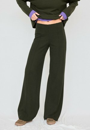 Personne portant un pantalon large vert foncé, un haut assorti et des chaussures marron, levant le haut pour montrer un sous-vêtement violet.