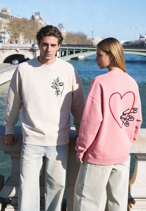 Homme en sweat crème avec motif abstrait visage et feuilles, femme en sweat rose avec cœur et épingles de sûreté, debout près de la rivière et du pont.