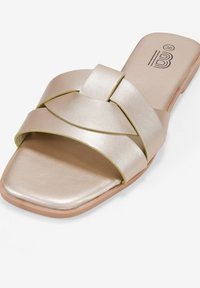 Sandalo scivolato in oro metallico con punta quadrata, presenta due cinturini incrociati e una suola piatta. Texture liscia con una leggera lucentezza.