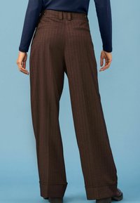 Inimene kannab kõrge vöökohaga pruuni triibulisi laiad jalad pükse, mille allääred on voltitud, ning tumesinist pikkade varrukatega ülikonda, seistes sinise taustaga.