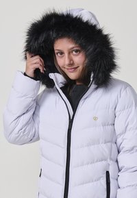 Chaqueta acolchada blanca con capucha de piel sintética negra. Incluye cierre de cremallera, textura acolchada y un pequeño emblema dorado en el pecho. Diseño cálido y ligero.