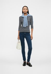 Ριγέ μακριμάνικο μπλουζάκι με ένα ανοιχτό μπλε πουλόβερ δεμένο γύρω από το λαιμό, σε συνδυασμό με σκούρους στενούς τζιν και μαύρα παπούτσια, κρατώντας μία λευκή τσάντα.