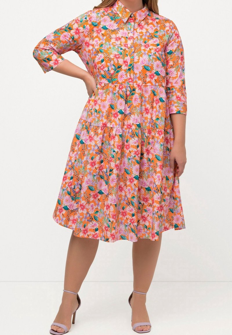 Femme portant une robe fleurie boutonnée, longueur genou, manches trois-quarts et talons à bride à la cheville, debout avec une main sur la hanche.