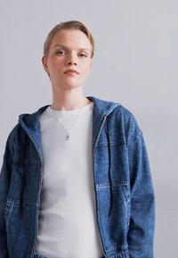 Denim zip-jakke med hætte, lys blå farve, med to frontlommer. Bæres over en hvid ribbet langærmet top.