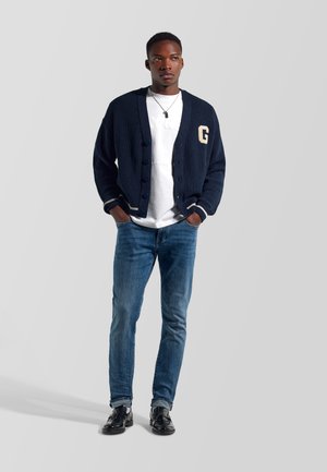 Hombre con un cárdigan azul marino con la letra G, camiseta blanca, vaqueros azules y zapatos negros, de pie con las manos en los bolsillos.