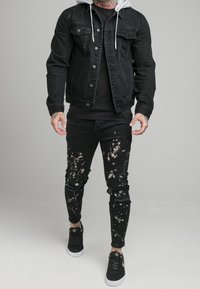 Man in een zwart denim jack over een zwarte hoodie, zwarte gespatte jeans en zwarte sneakers met witte zolen, staande op een lichtgrijze vloer.