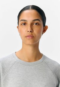 Lys grå sweatshirt med en glat tekstur og raglanærmer, der har en rund halsudskæring, båret af en person med glat sort hår.