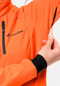 Orange jacka med dragkedjeficka, svarta ärmslut och justerbar handled. Tillverkarens logotyp synlig på framsidan; textil med struktur synlig.