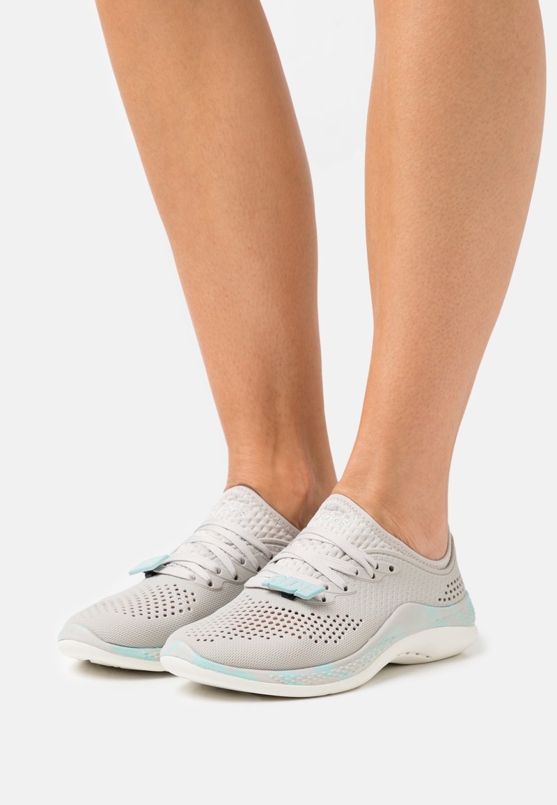 Personne portant des baskets en mesh respirant gris clair avec des semelles blanches et des accents bleu clair sur un fond blanc.