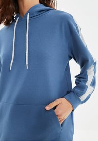 Mujer vistiendo una sudadera azul con cordones blancos y rayas gris en las mangas, con la mano dentro del bolsillo frontal.