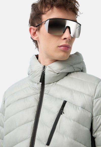 Gepolsterte hellgraue Jacke mit hohem Kragen, schwarzen Reißverschlüssen und Brusttasche. Das Model trägt reflektierende übergroße Sonnenbrillen.