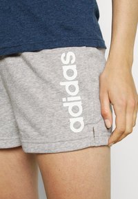 Närbild av en person som bär grå Adidas-shorts med vit logotyp på låret och en mörkblå skjorta, med den högra handen vilande bredvid shortsen.