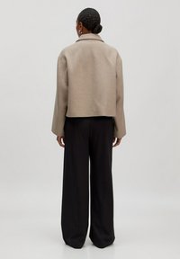 Vrouw met een knot, gekleed in een beige cropped jas en zwarte wijde broek, staande met haar rug naar de camera tegen een effen achtergrond.