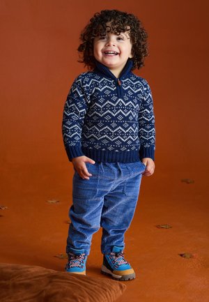 Kleinkind mit lockigem Haar lächelt, trägt einen blau gemusterten Pullover, blaue Cordhosen und bunte Wanderschuhe auf einem orangefarbenen Boden.