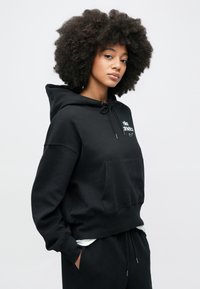 Μαύρο φούτερ με κουκούλα, μπροστινή τσέπη, κορδόνι στο κλείσιμο και λογότυπο σε λευκό χρώμα. Μαλακό ύφασμα με άνετη εφαρμογή.