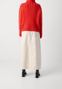 Person iført en rød strikket sweater, lang beige nederdel med baglommer og sorte hælede ankelsko, stående foran en ensfarvet hvid baggrund.