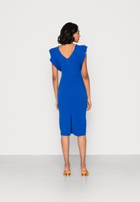 Vestido azul com mangas onduladas, costas em V, de comprimento até ao joelho, com um design ajustado, apresentando um fecho traseiro e uma fenda central nas costas. Sapatos de salto laranja.