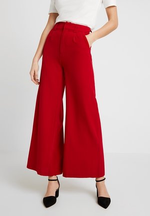 Kobieta ubrana w czerwone spodnie z wysokim stanem i szerokimi nogawkami, biały krótki rękaw, czarne szpilki na kostce, stojąca z jedną ręką w kieszeni.