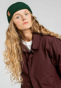 Zielona dzianinowa czapka z logo ze skóry, zestawiona z bordową kurtką z kołnierzem na białej koszulce. Modelka ma długie, faliste włosy.
