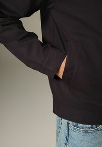 Schwarze Jacke mit strukturiertem Design, ausgestattet mit einer versteckten Tasche und detaillierten Nähten. Kombiniert mit blauer Jeans, die einen lässigen Look präsentieren.