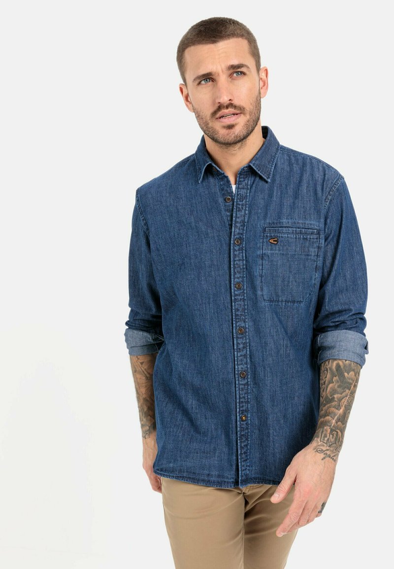 Denimskjorte i mørkeblå med knaplukning foran, brystlomme og opkrøllede ærmer. Har en klassisk krave og diskrete syninger.
