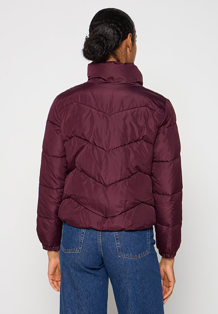 Person mit dunklem Haar im Dutt, der eine gesteppte kastanienbraune Jacke und blaue Jeans trägt und mit dem Rücken zu einem einfarbigen weißen Hintergrund steht.