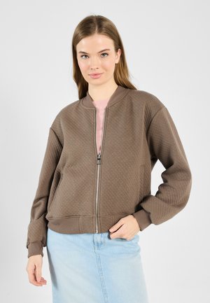 Veste bomber matelassée marron avec fermeture éclair sur le devant, poignets et ourlet côtelés. Portée sur un haut roses clair, avec une jupe en jean bleu clair décontractée.
